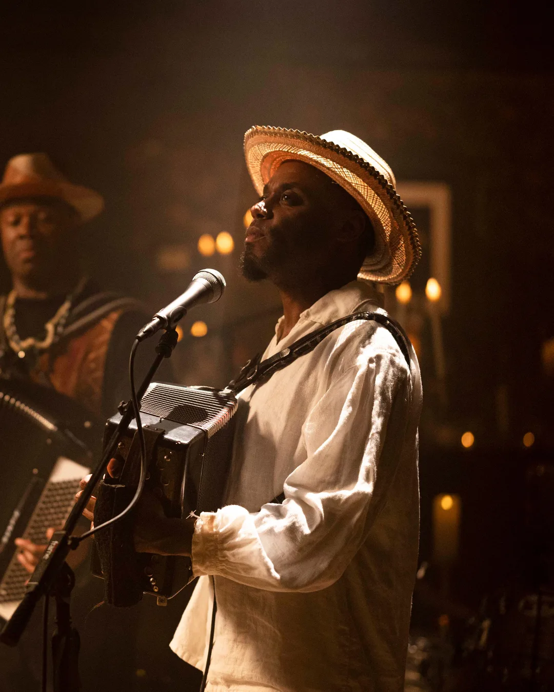 A musician in a straw hat and white shirt sings at a microphone while band members playing guitar, drums, and other…