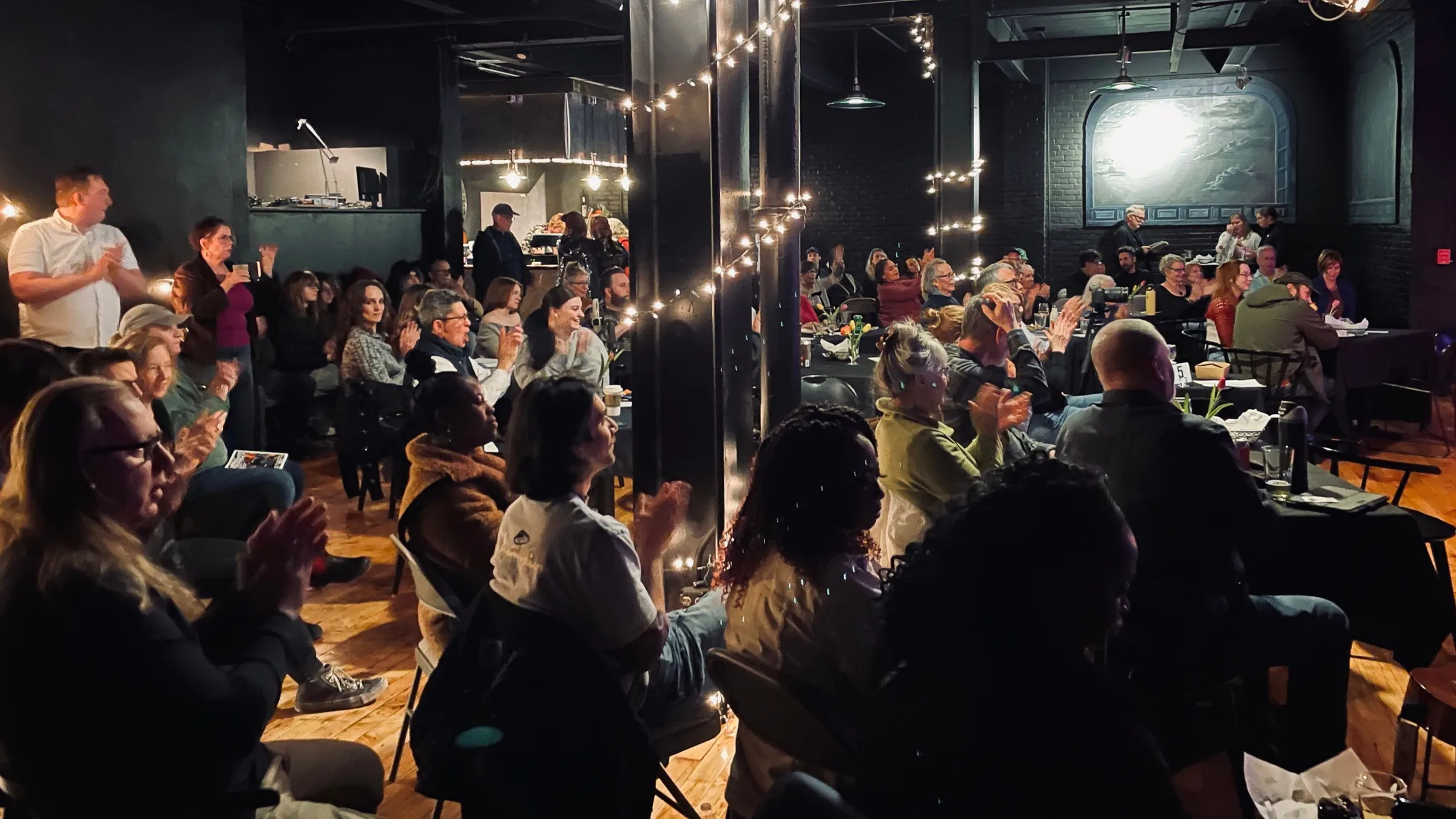 An audience at tables and in rows, applauding, among black columns wound with string lights