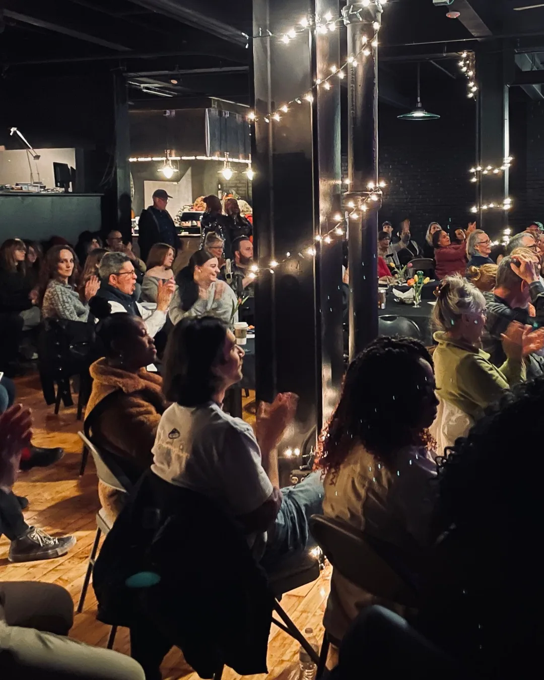 An audience at tables and in rows, applauding, among black columns wound with string lights