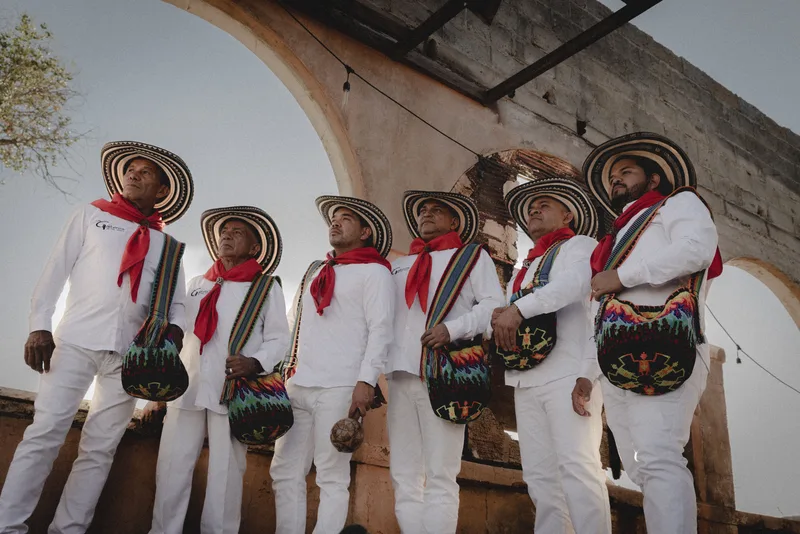 Six musicians in white traditional outfits, wide-brimmed hats, red neck scarves, and colorful woven bags perform outdoors…