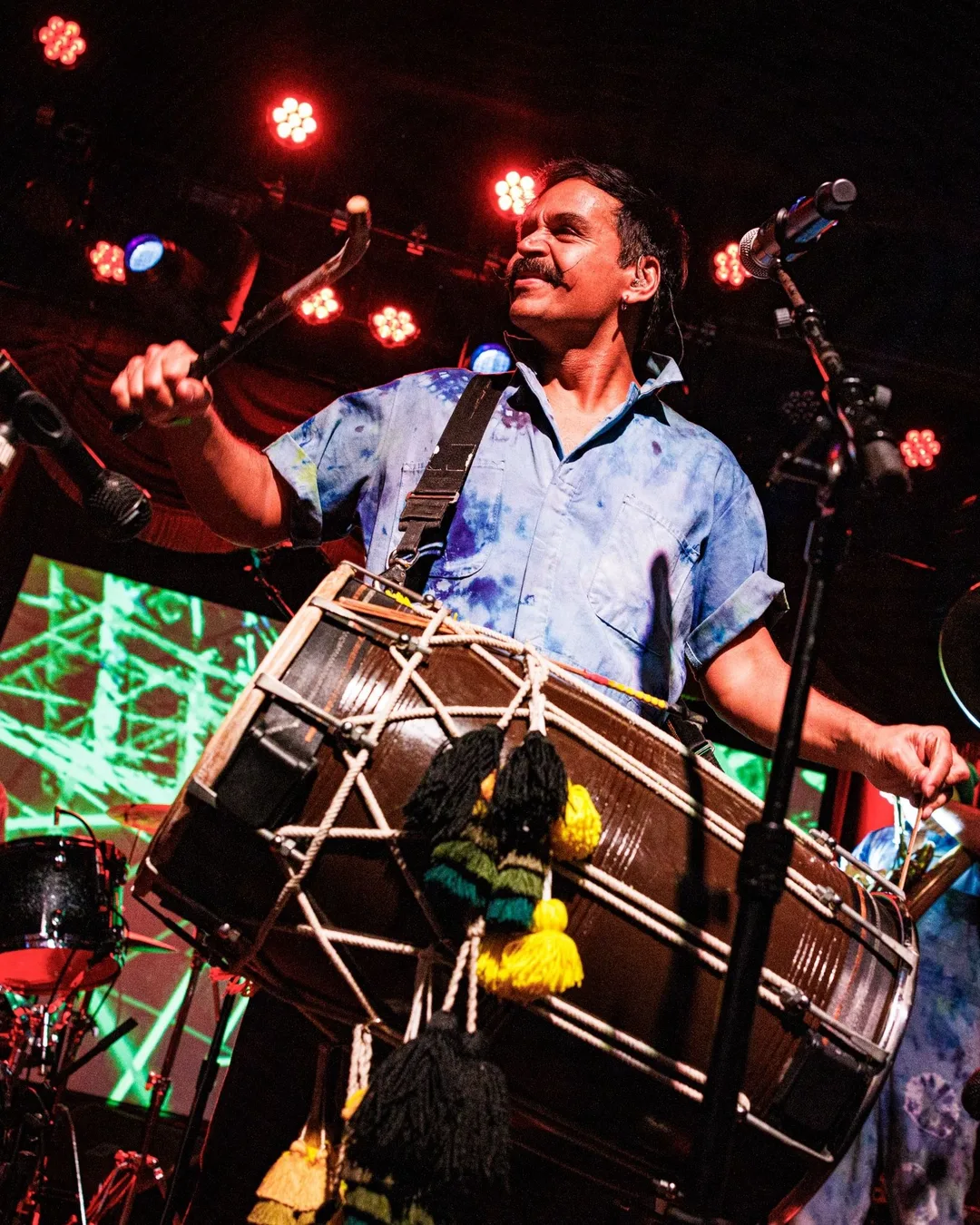 A musician in a tie-dye shirt plays drums on stage under red and blue stage lights, with band members and a green-lit…