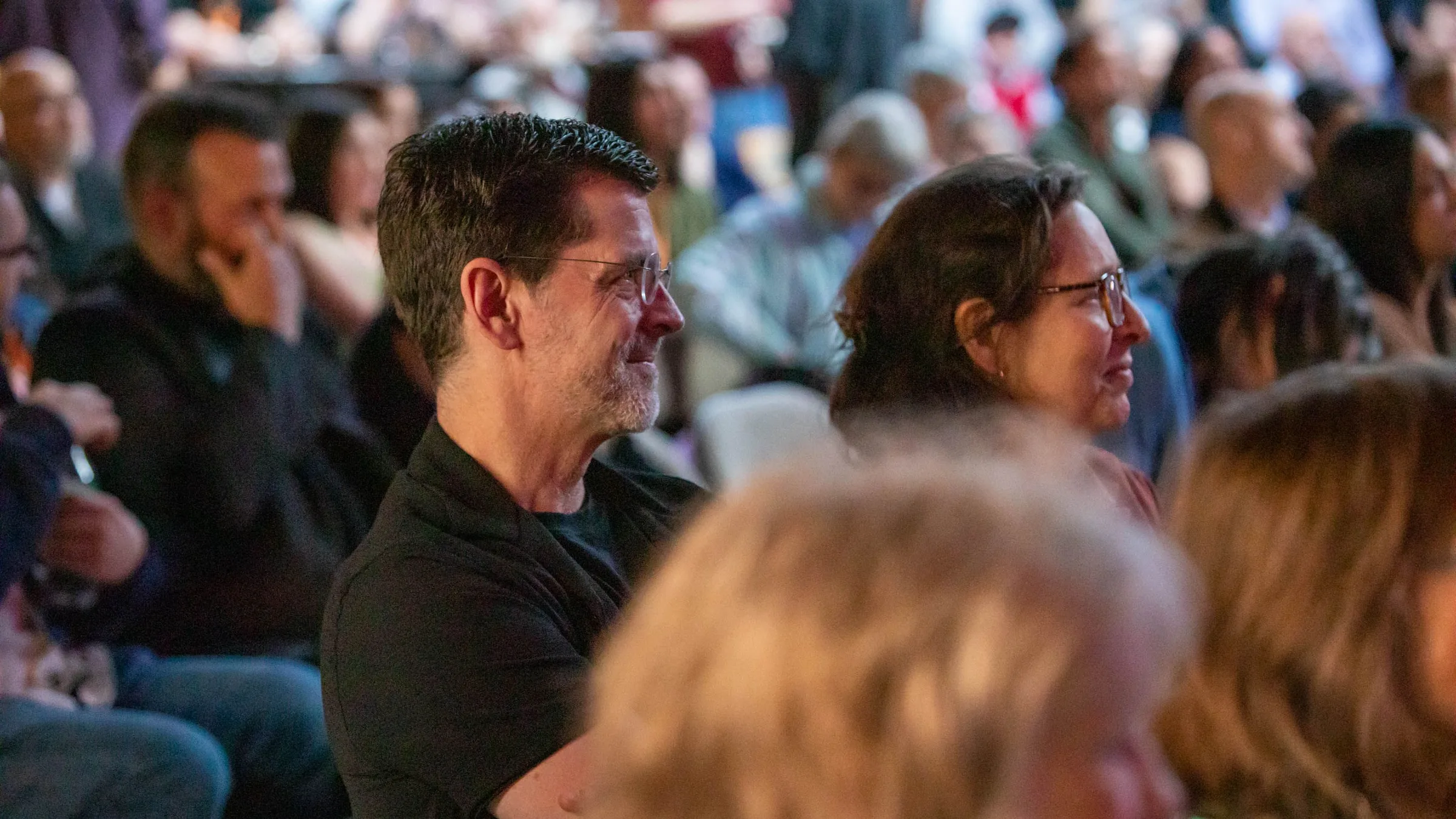 Two members of an audience smiling as they listen, the room full behind them
