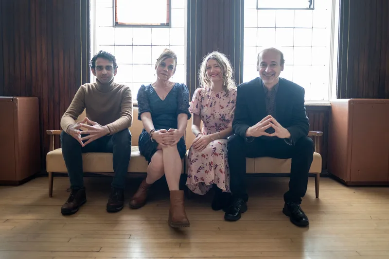 Four musicians sit side by side on a tan couch in front of large windows, smiling at the camera in a bright room with wood…