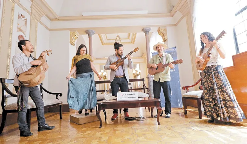 Five musicians perform with acoustic guitars in an ornate cream and gold room with classical columns and wooden floors.
