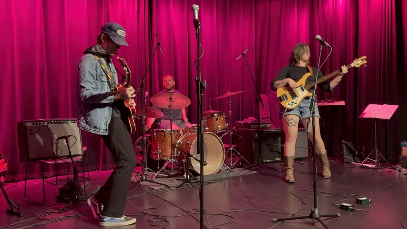 Four musicians perform on stage with bright magenta curtains behind them: a guitarist in a cap and denim shirt on the left…