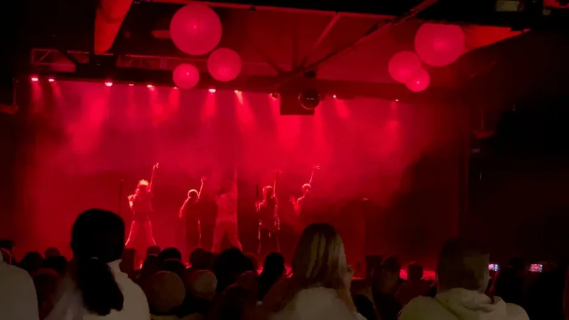Band performing on stage with red lighting and spherical fixtures while audience members watch from the floor.