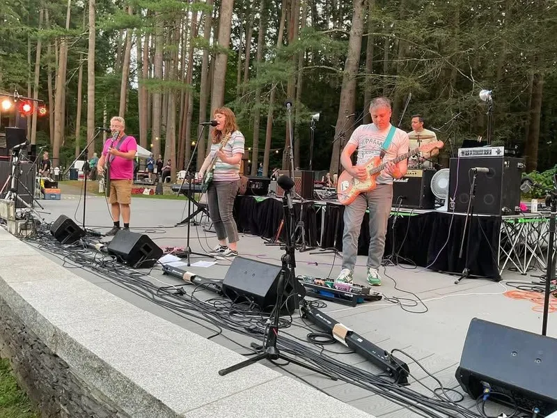 Live band performs outdoors among tall trees, with four musicians playing vocals, guitar, and drums on a concrete stage…