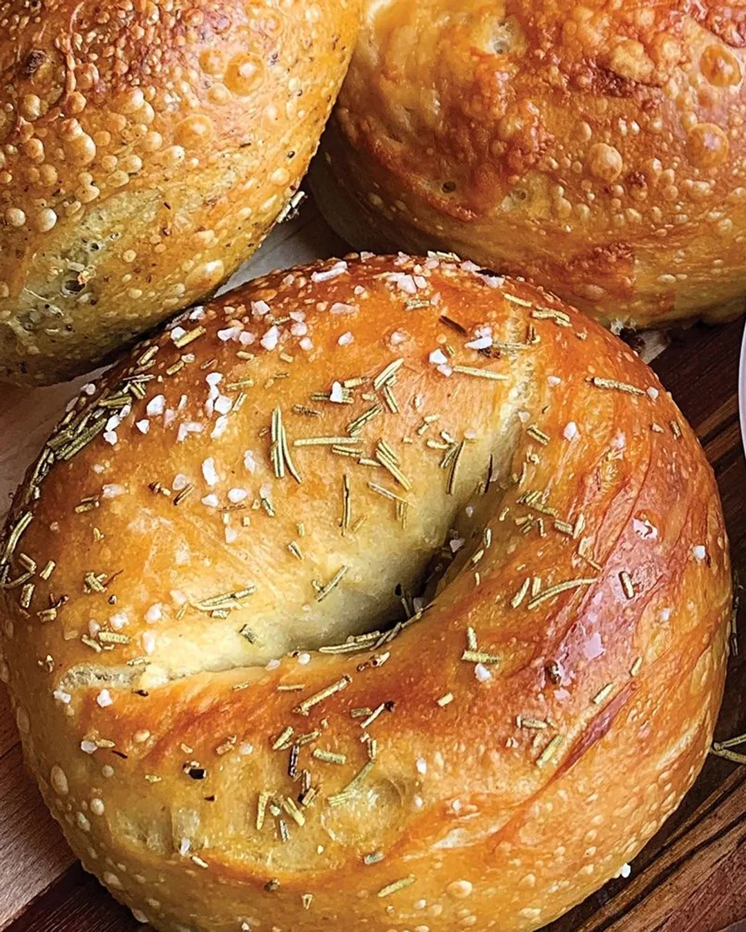 Sourdough bagels on a wooden board — one topped with rosemary and flaky salt, one with seeds — beside a tub of scallion cream cheese