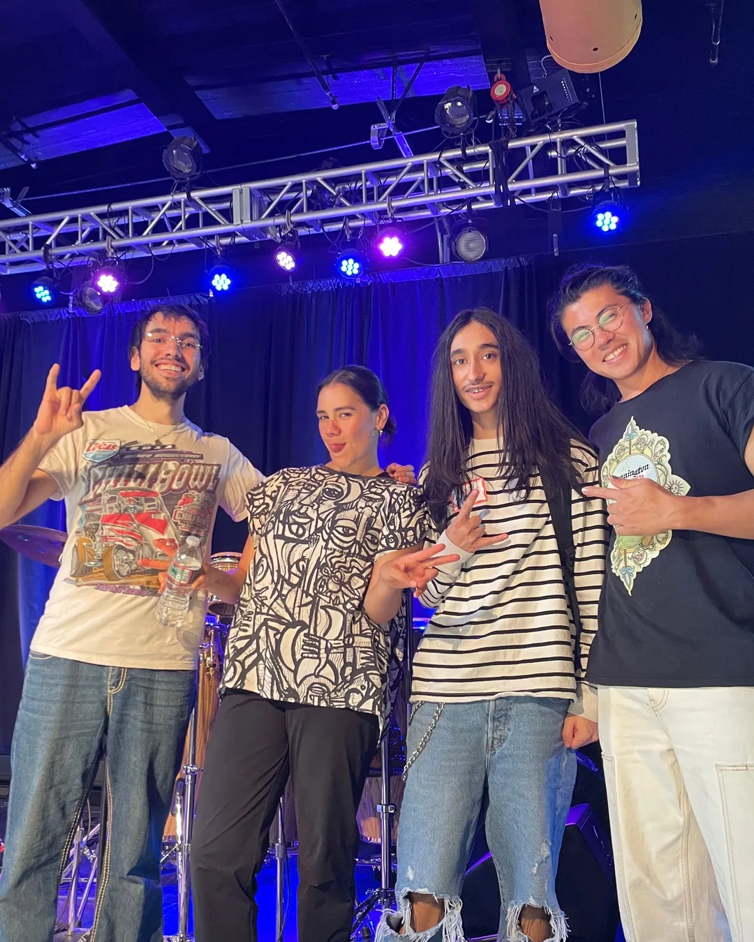 Four young people on stage under the lighting truss, grinning, one throwing a rock-and-roll sign