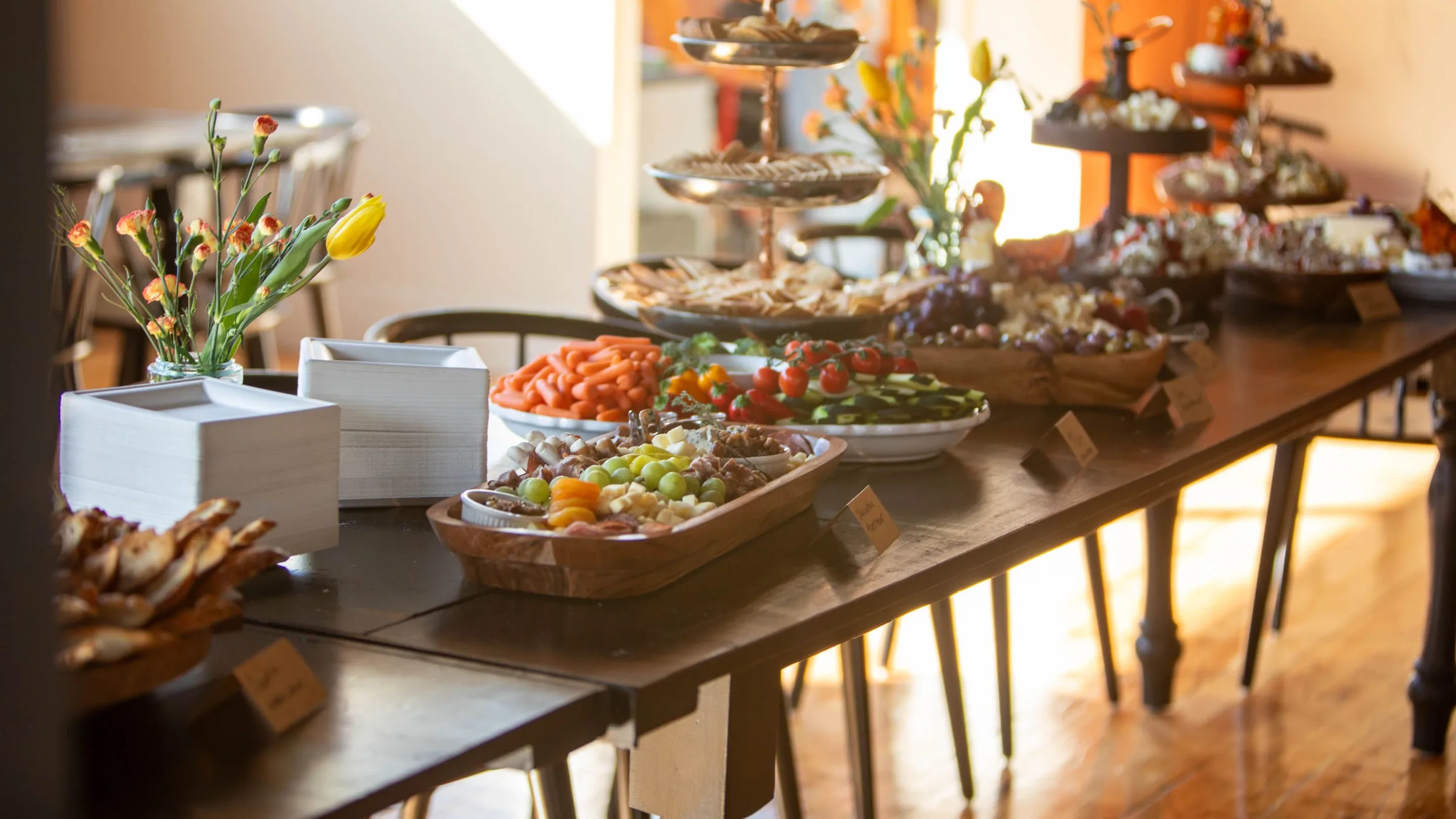 A buffet on long wooden tables in afternoon light: tiered stands, fruit, crudités, crackers and small labelled cards