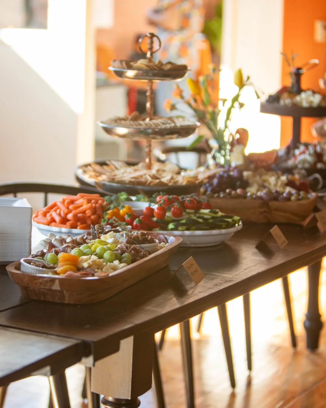 A buffet on long wooden tables in afternoon light: tiered stands, fruit, crudités, crackers and small labelled cards
