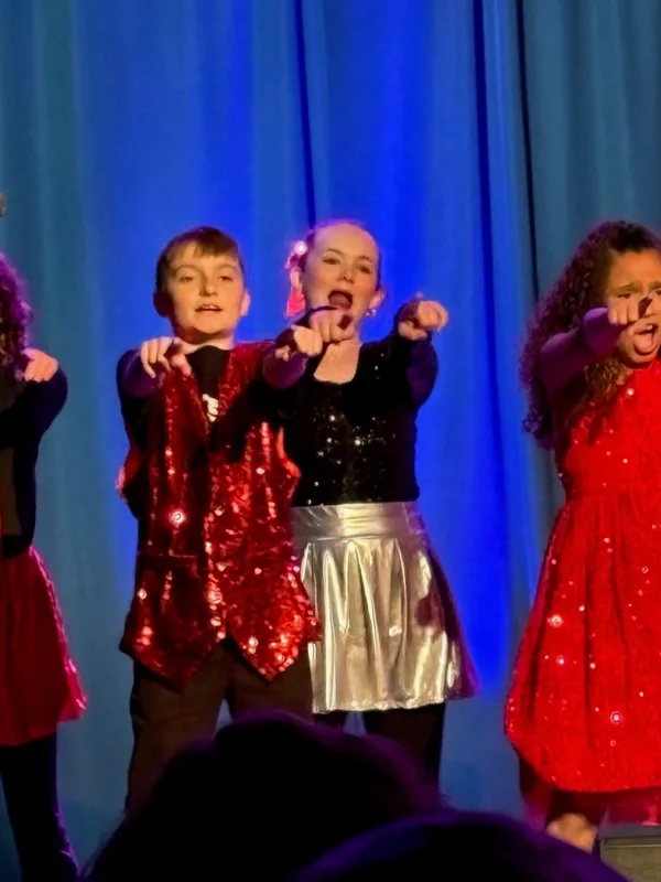 Young performers in sequined red and black outfits sing and dance together on stage under blue stage lighting.