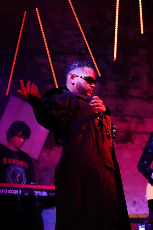 A performer wearing sunglasses and a black jacket gestures on stage beneath vertical red neon lights and purple ambient…