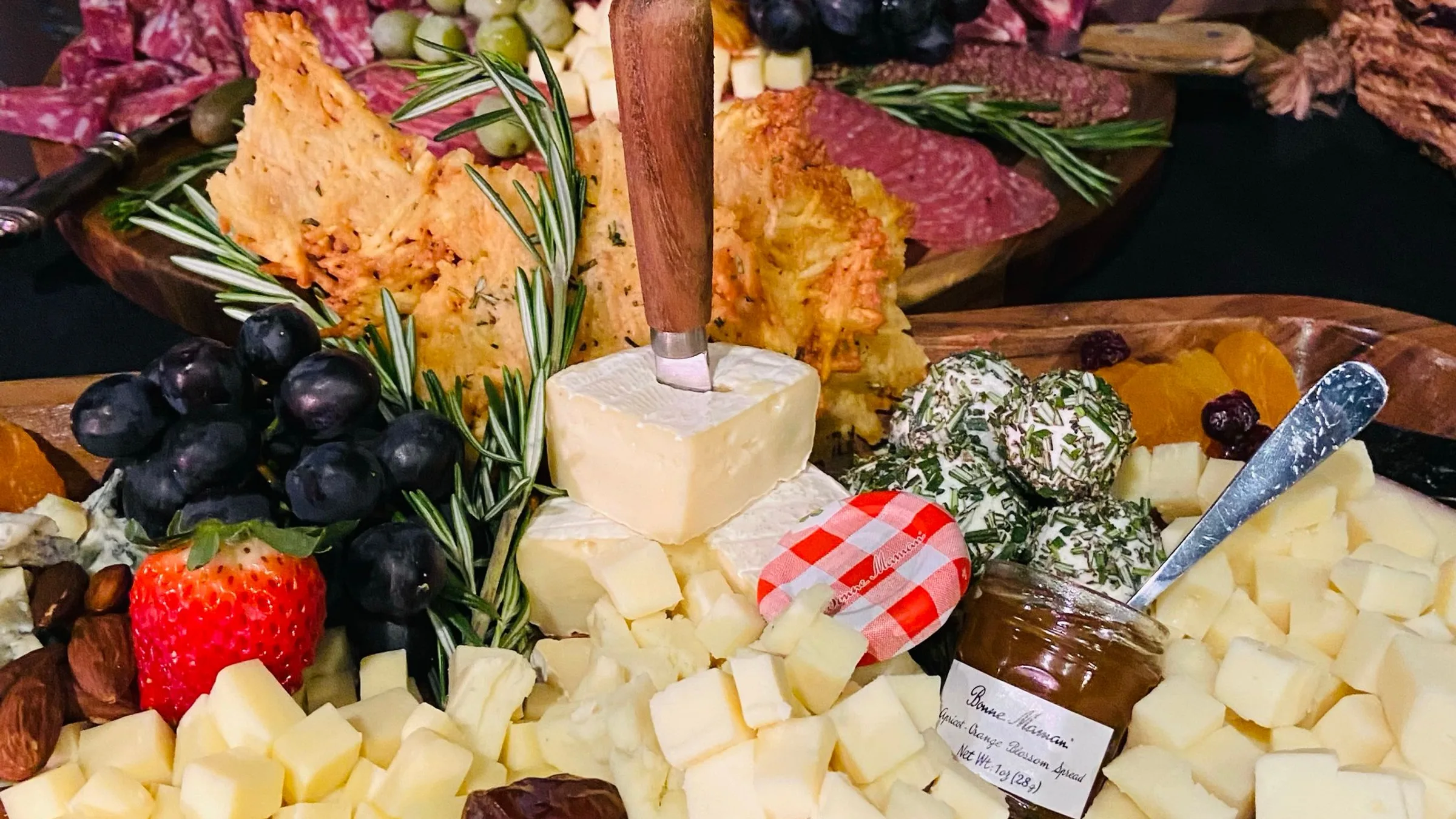 A wooden board of cubed cheeses, cured meats, grapes, crisps and a jar of preserves against a black brick wall