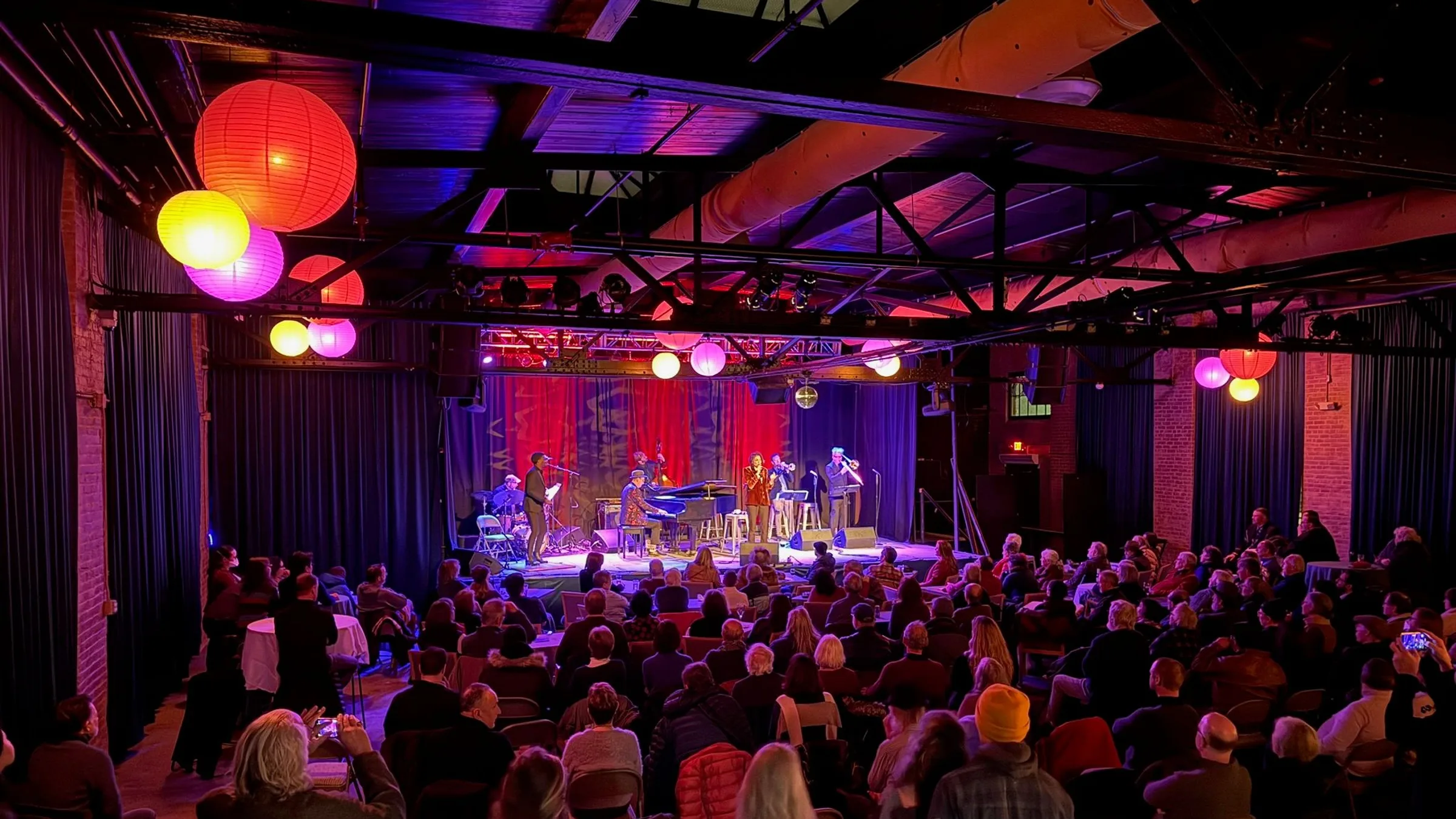 The Soundstage from the back of the room: rows of seated guests facing a lit stage under paper lanterns and steel trusses