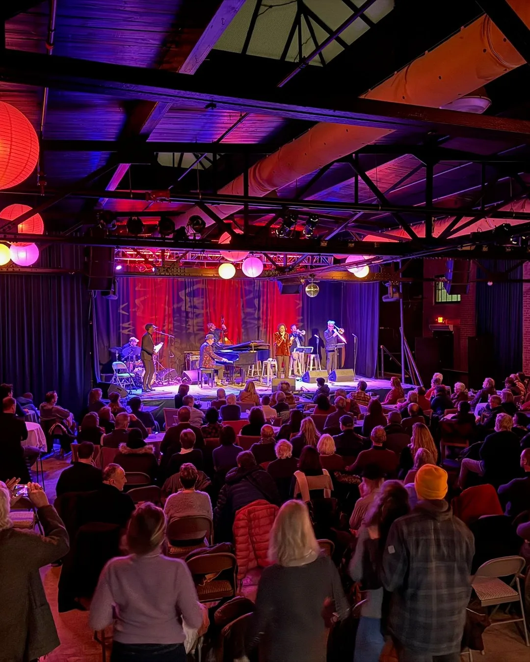 The Soundstage from the back of the room: rows of seated guests facing a lit stage under paper lanterns and steel trusses