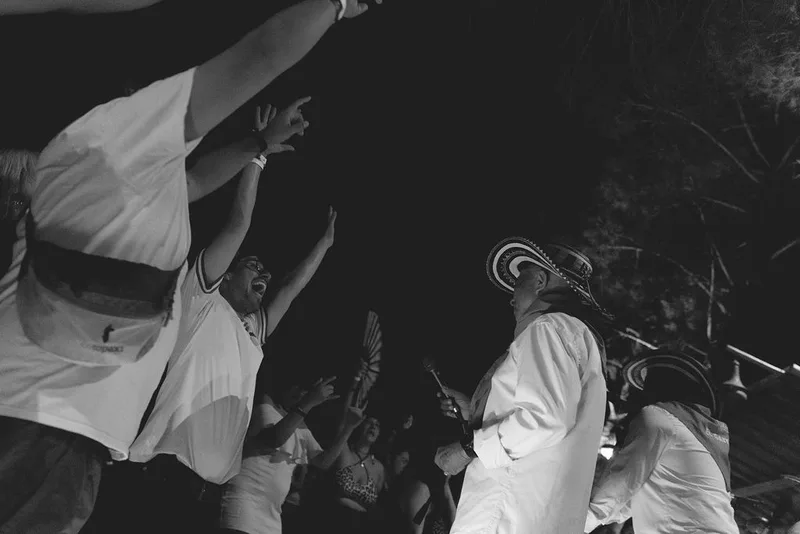 Performer in cowboy hat and white shirt plays guitar on stage while audience members in light-colored clothing dance with…