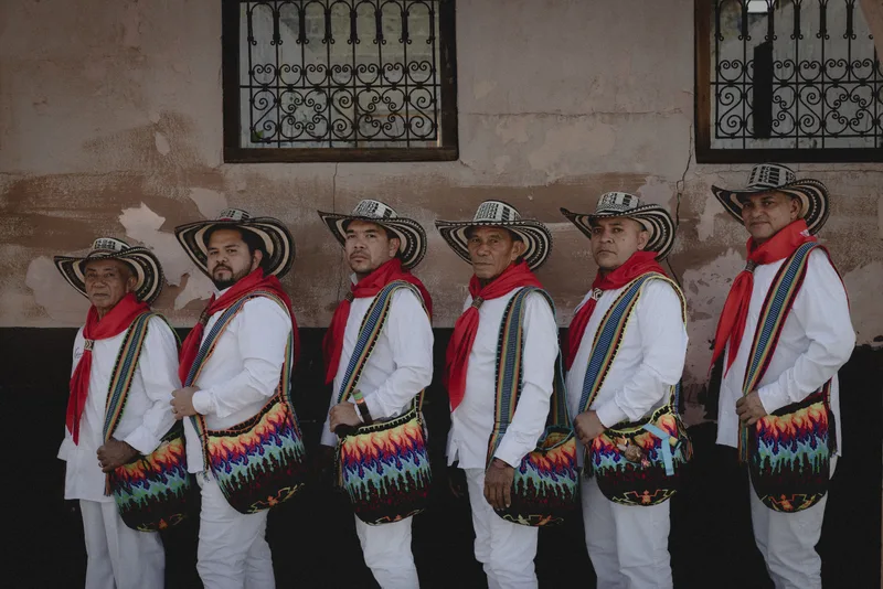 Six musicians in traditional Colombian attire stand in a line wearing white shirts, red scarves, woven hats, and colorful…