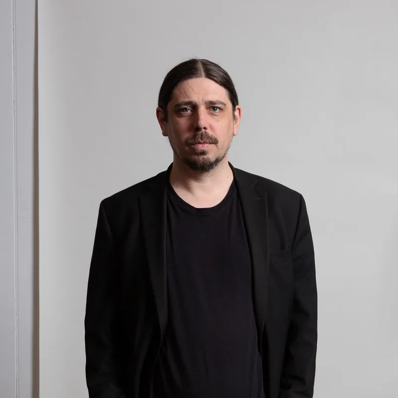 Man with dark hair and facial hair wearing a black blazer and black t-shirt against a neutral gray background.