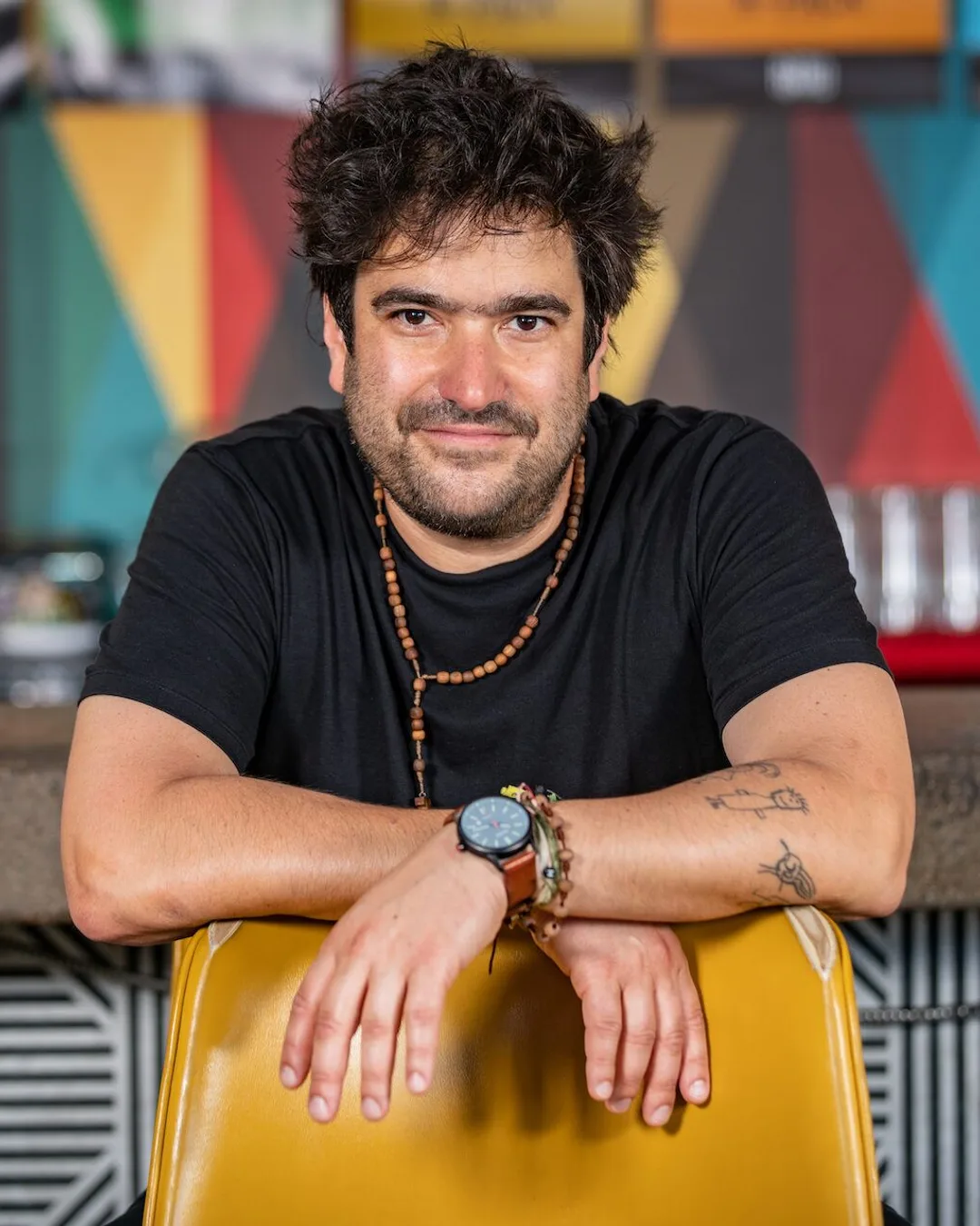 Man with dark hair and beard wearing black shirt and beaded necklace leans against yellow chair in front of colorful…