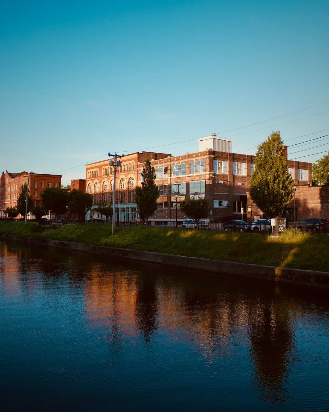A row of historic red brick buildings sits along a calm canal under clear blue sky, with utility poles and green vegetation…