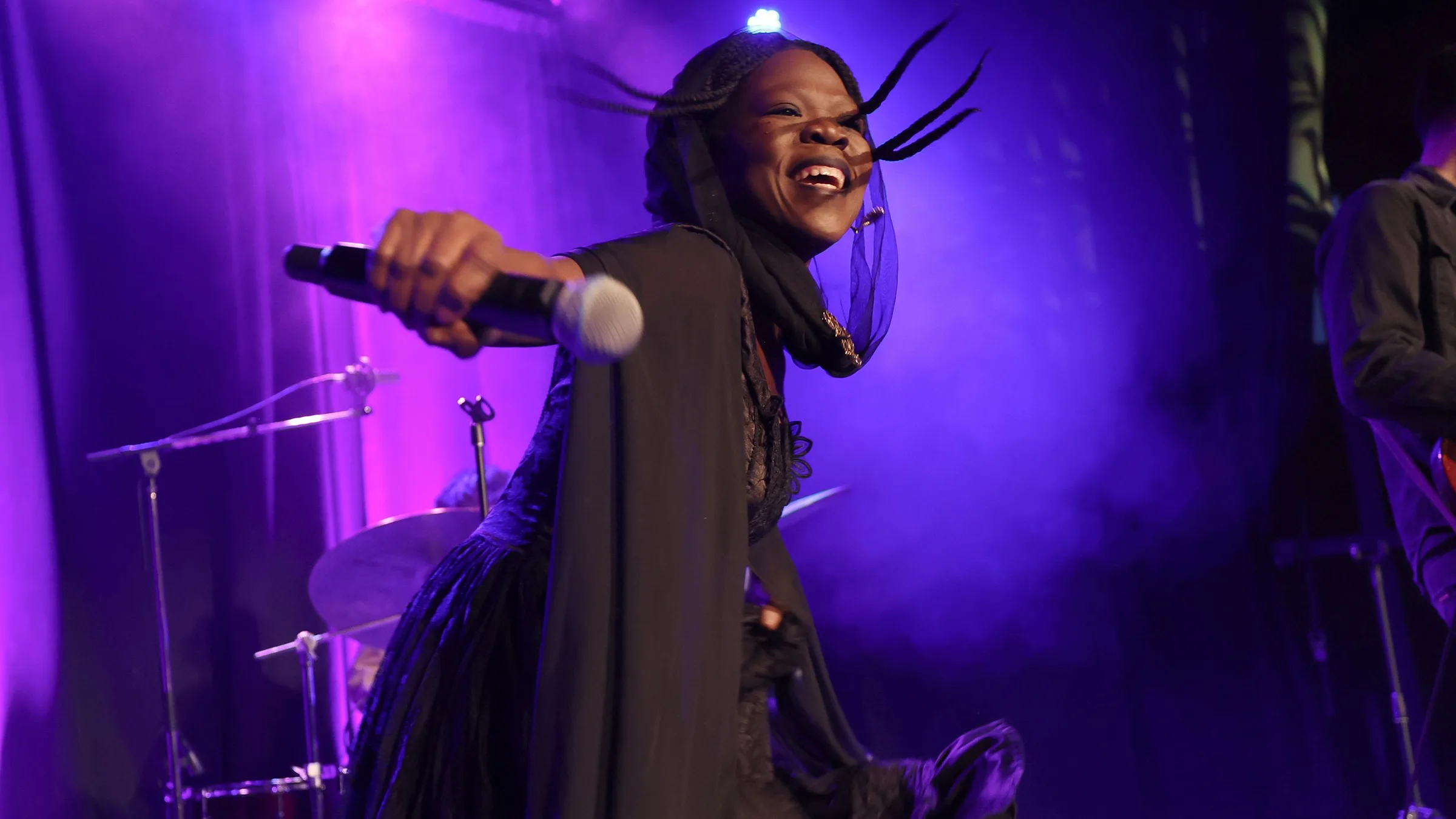 Moonlight Benjamin, in a black veil and gown, laughing and holding a microphone out to the crowd under violet light
