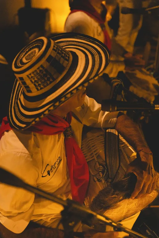Musician wearing a patterned straw hat and red sash plays guitar under warm amber stage lighting.