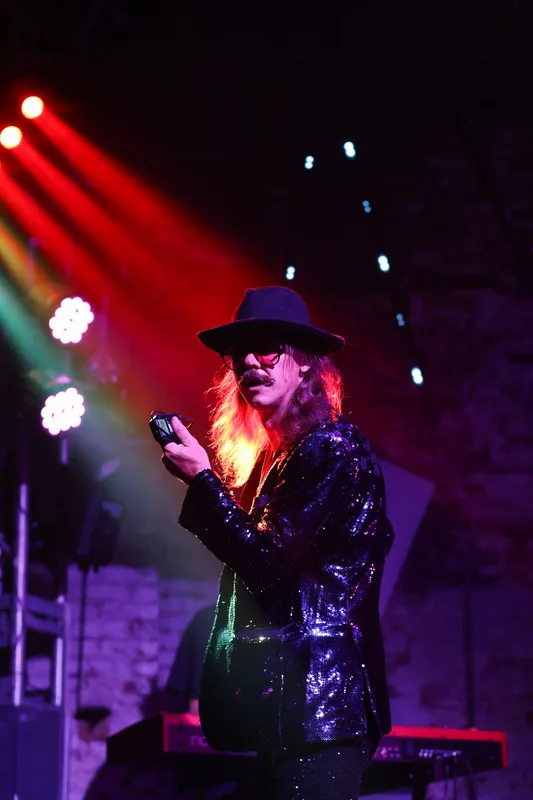 Performer in a sequined jacket and wide-brimmed hat holds a microphone on stage under red and blue stage lighting.