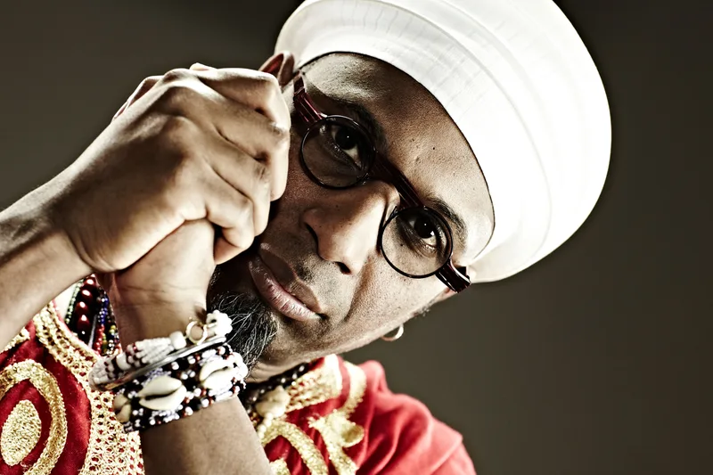 Performer wearing a white wide-brimmed hat, dark sunglasses, and ornate red and gold jacket with beaded accessories…