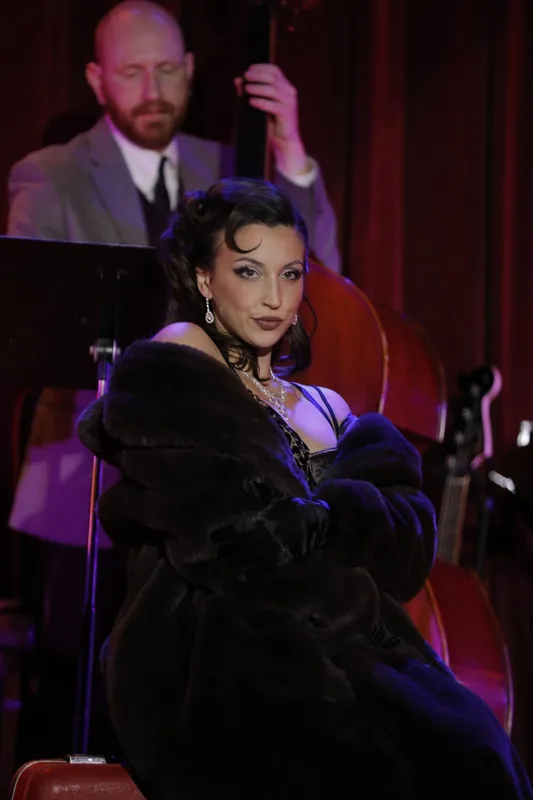 A woman in a black velvet jacket and jewelry sings at a microphone while a man in a suit plays upright bass behind her…