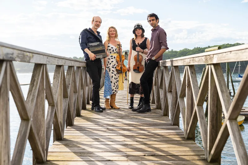 Four musicians stand on a wooden pier holding an accordion and two violins, with water and hills visible behind them.