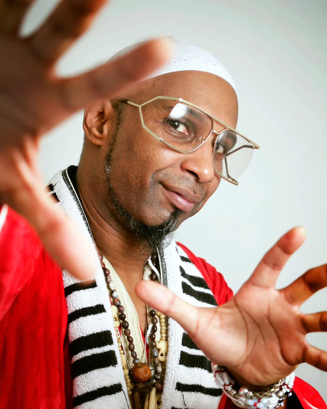 A man wearing glasses, a white headwrap, red jacket, and striped shirt gestures expressively with both hands against a…