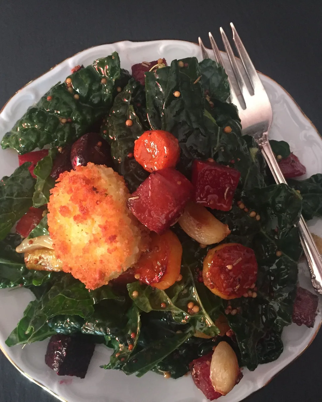 A plate of green kale salad with roasted beets, crispy orange croquette, and mustard seeds, served with a fork on dark…