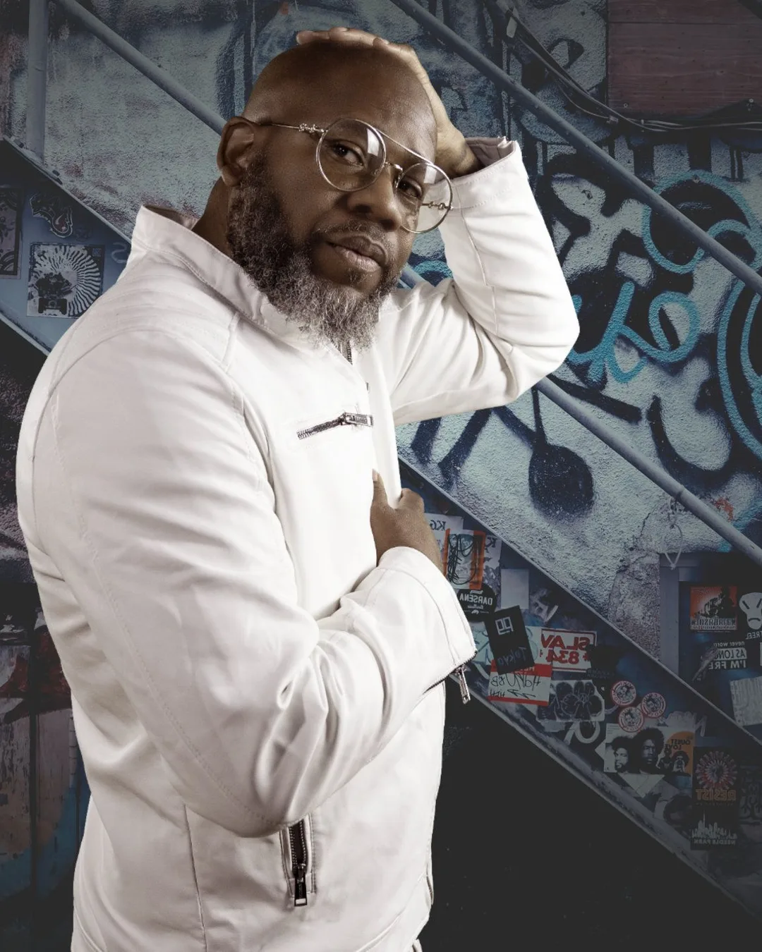 Man wearing glasses and white hoodie poses against industrial staircase with graffiti-covered walls.