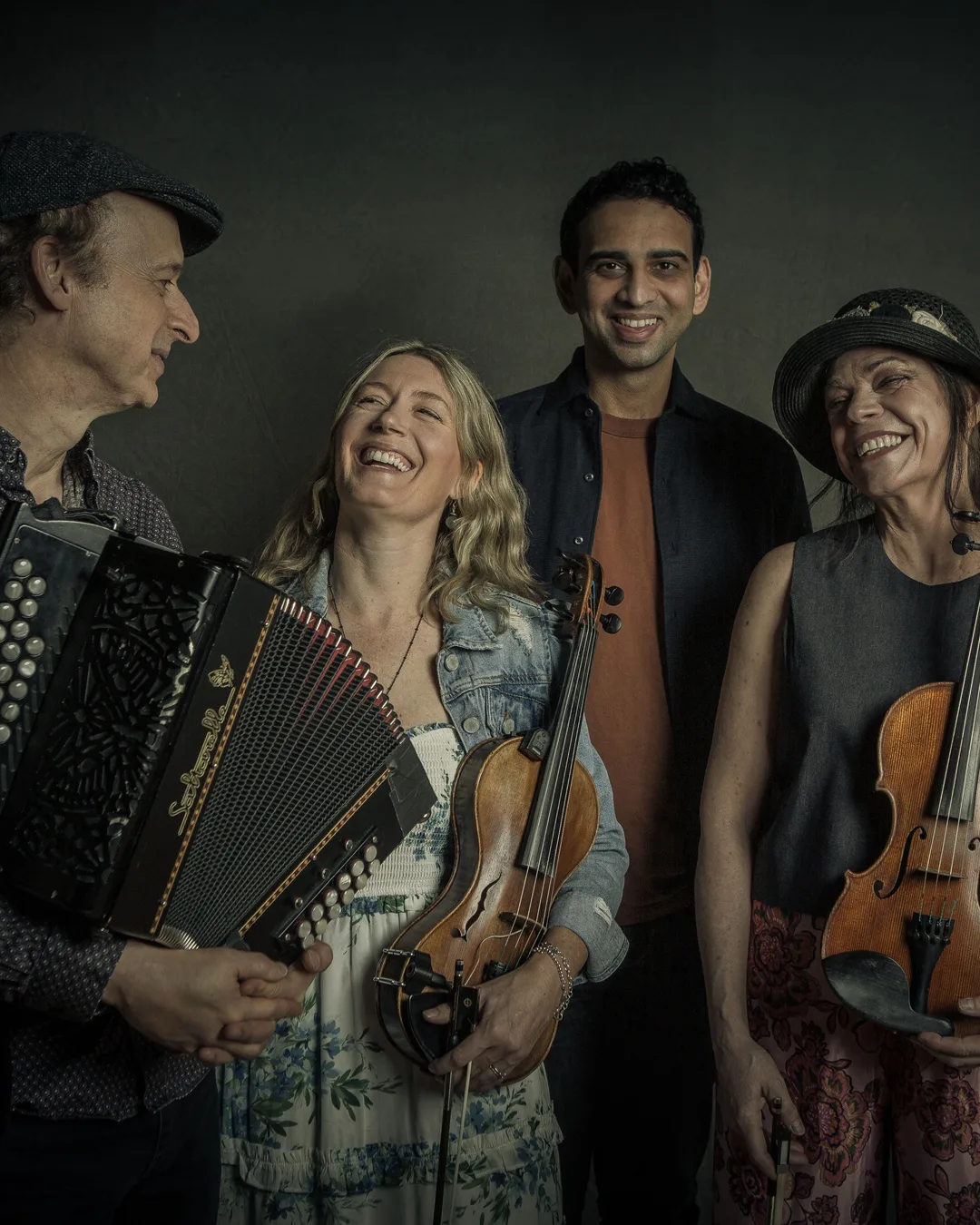 Four smiling musicians hold an accordion and two violins against a dark green background.
