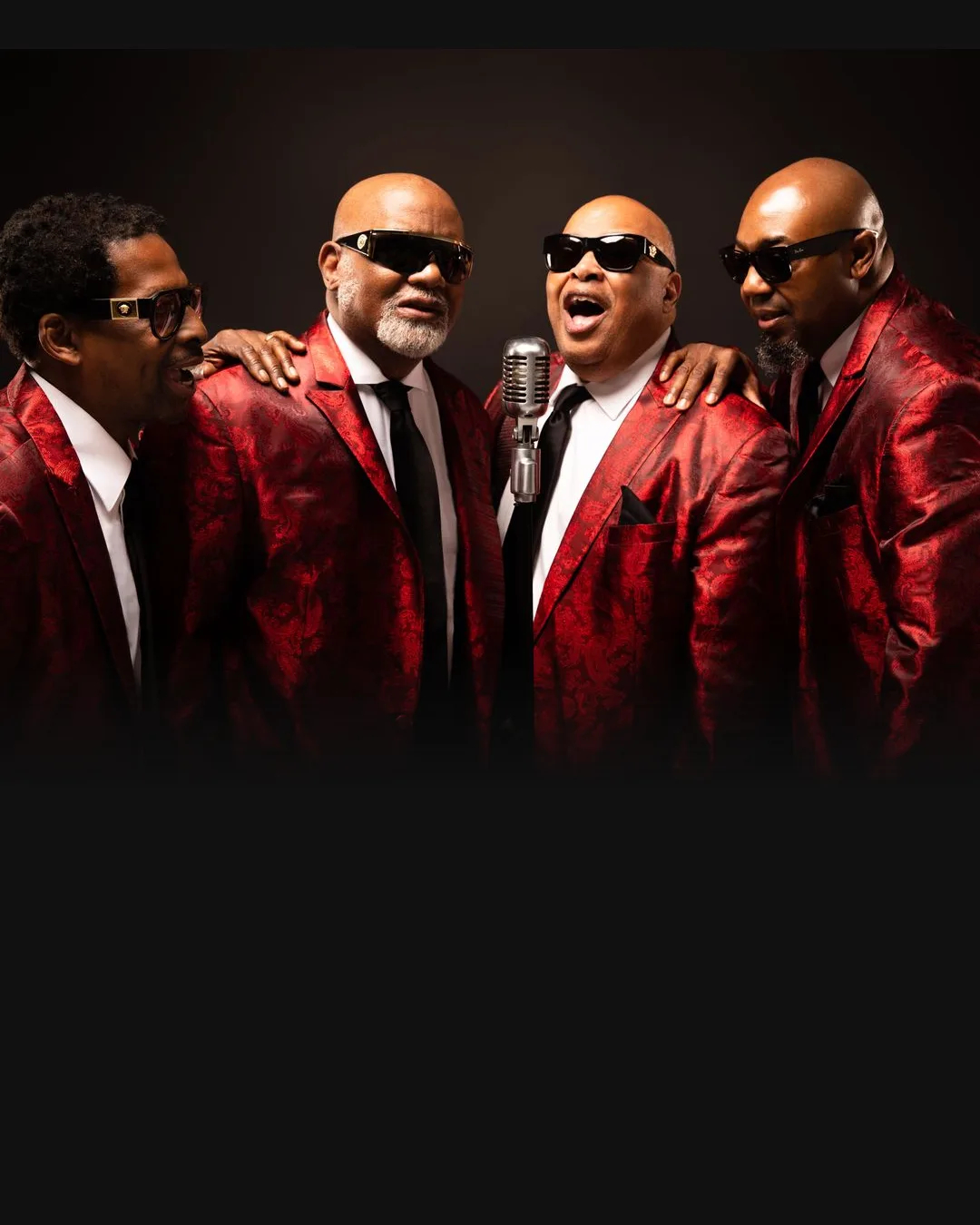 Four men in red patterned jackets and sunglasses perform around a vintage microphone against a black background.