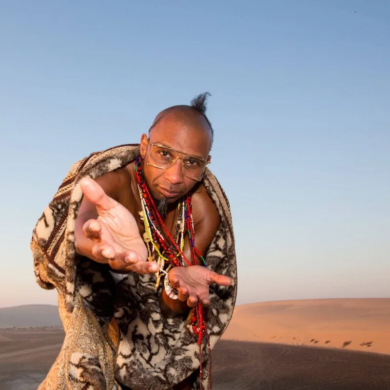 Man wearing patterned jacket and beaded necklaces gestures toward camera against a desert landscape backdrop.