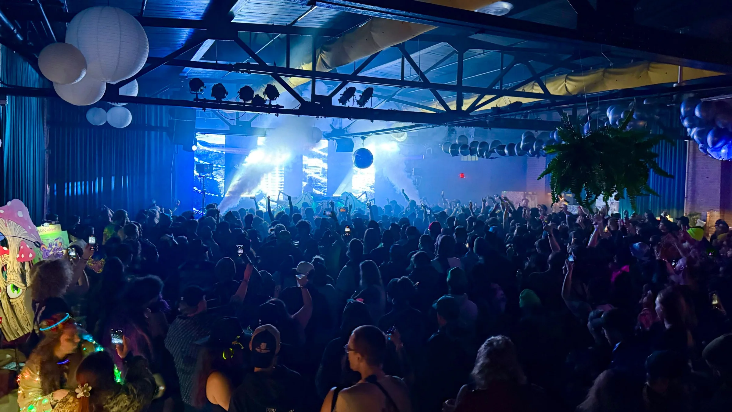 A packed standing crowd with hands in the air facing a stage washed in white light and haze