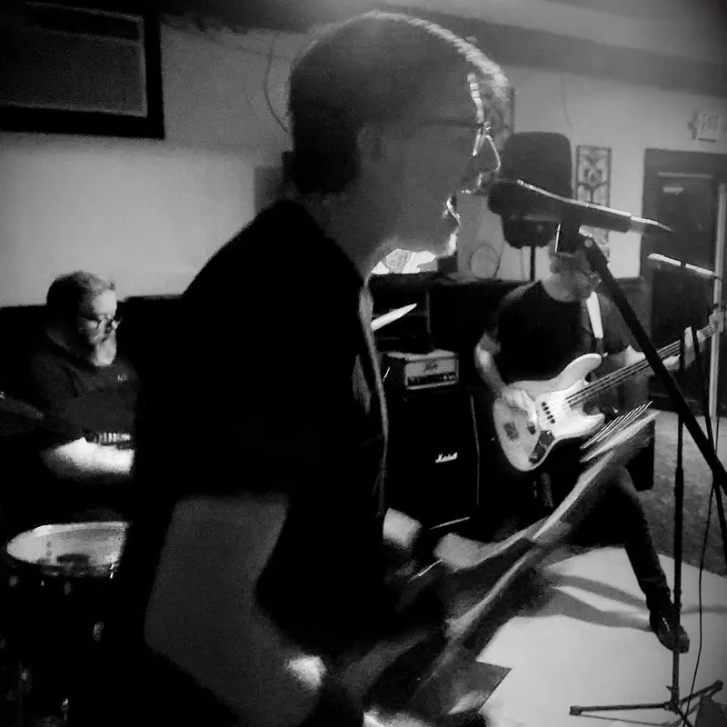 Band performing in an intimate indoor venue with a singer at the microphone, bass guitarist, drummer, and audience member…
