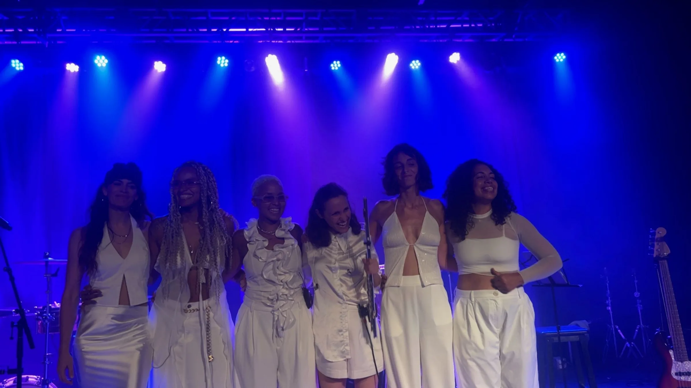 Six performers dressed in white, arm in arm, taking a bow under blue stage light