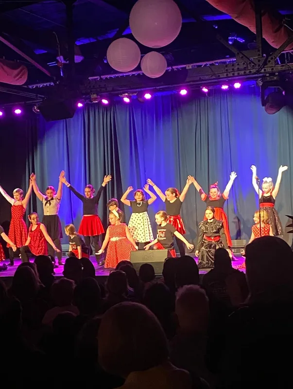 A group of young performers in vintage-style dresses dance on stage with raised arms under blue and pink stage lights, with…