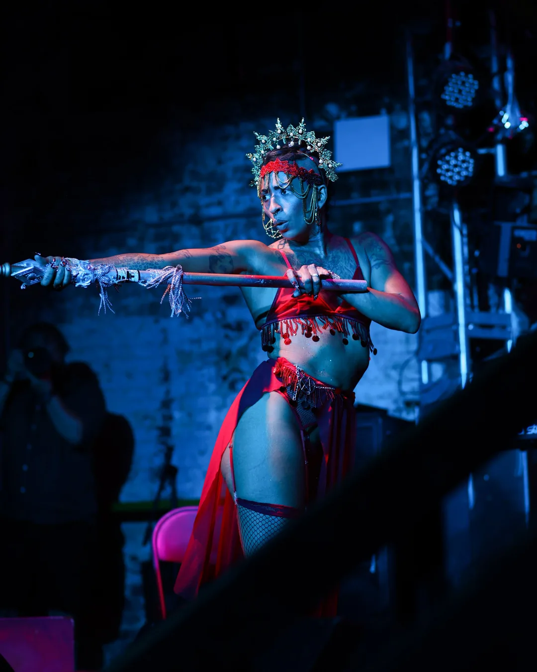 Performer in ornate silver crown and red costume holds a staff on stage under blue stage lighting with audience members…