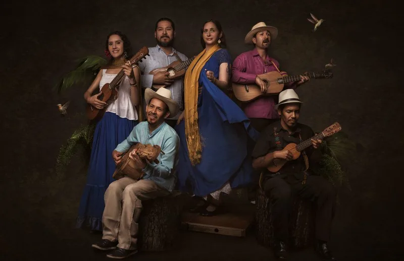 Six musicians holding acoustic guitars posed together against a dark background with green foliage and warm lighting.