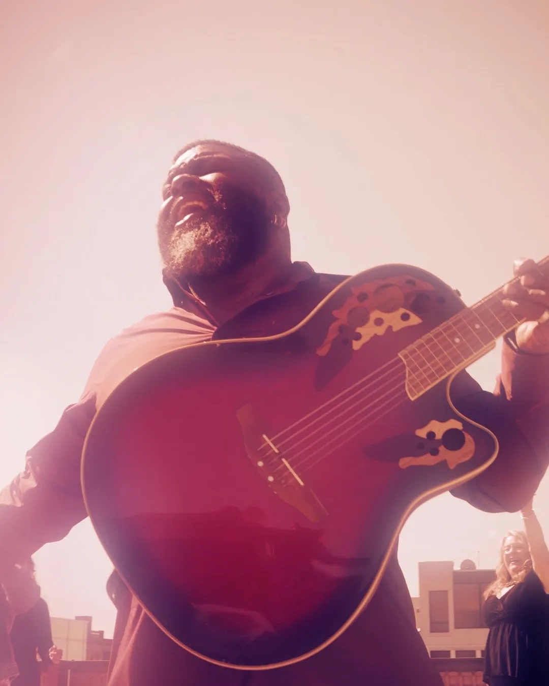 A bearded guitarist in a red hoodie plays an acoustic guitar while singing, bathed in warm golden light with band members…