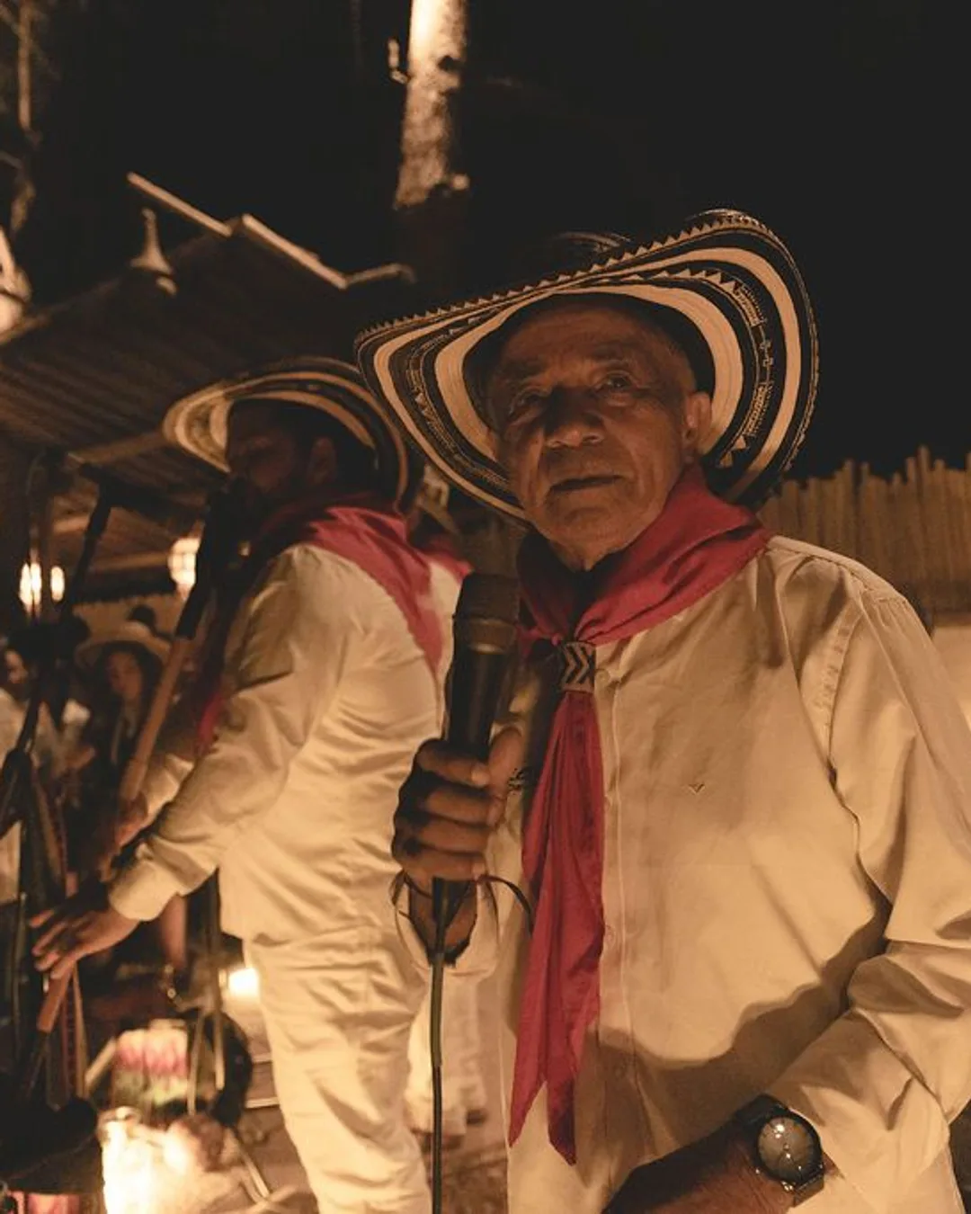 Man in wide-brimmed sombrero and cream-colored shirt singing at microphone with band members and guitars in warm stage…