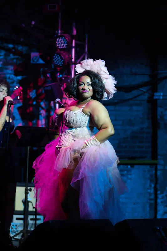 A performer in a white flower headpiece and pink tulle gown sings at a microphone under magenta and blue stage lighting.