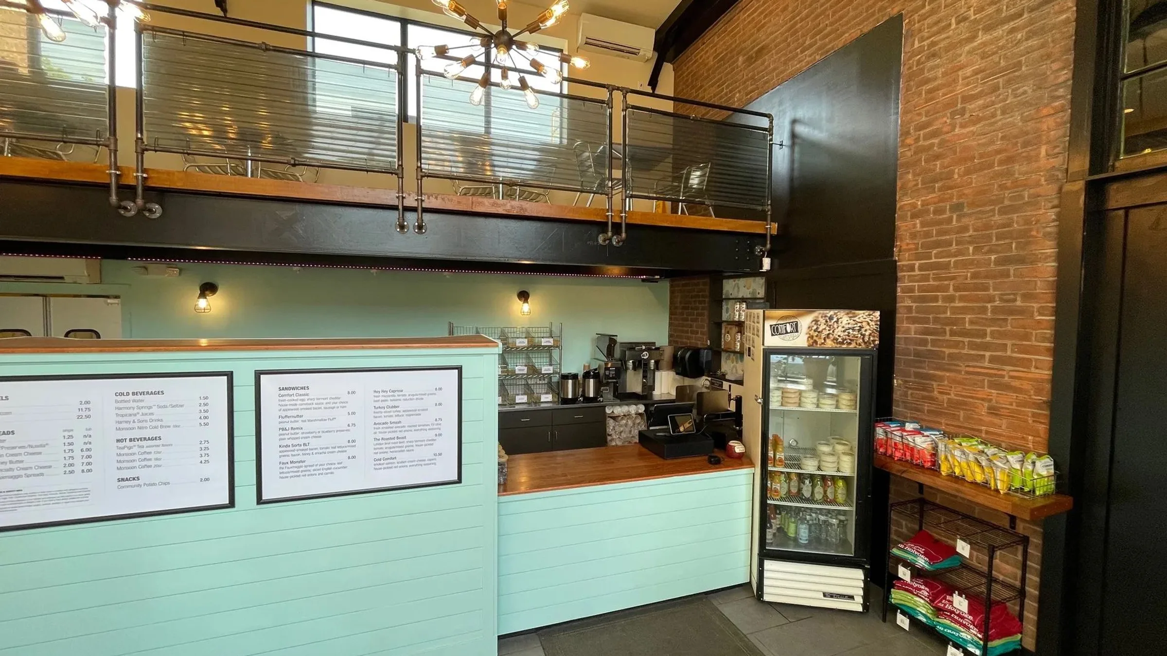 The Comfort Bagel counter: a mint-green panelled bar, menu boards, a drinks fridge and a snack rack against exposed brick, under a mezzanine rail and a sputnik chandelier
