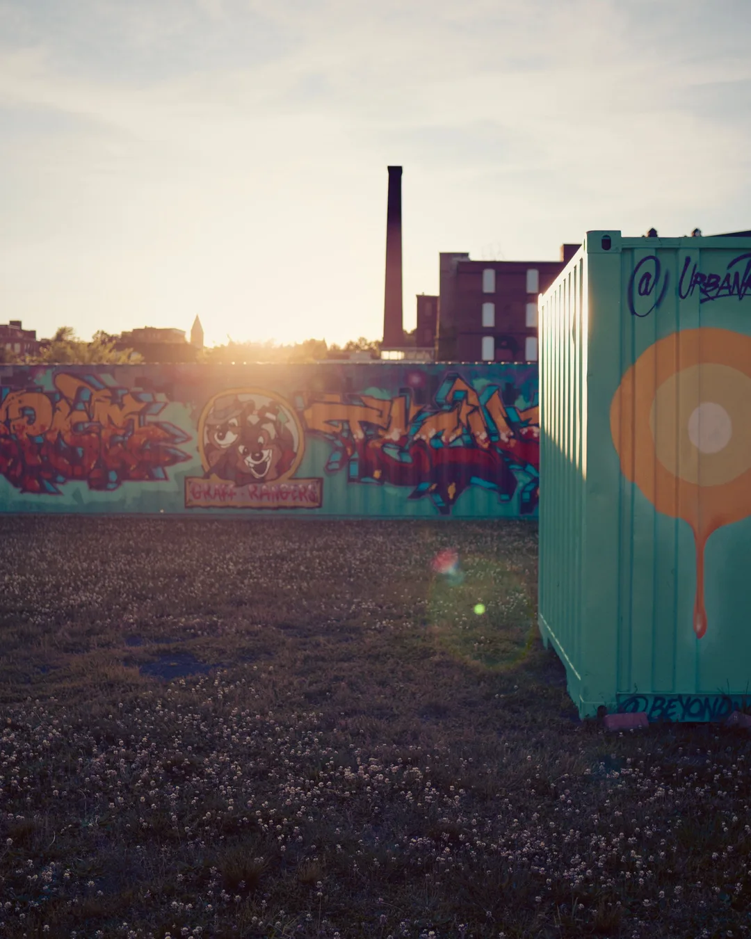 Outdoor venue with colorful graffiti-covered walls and shipping containers at sunset, industrial buildings and a smokestack…