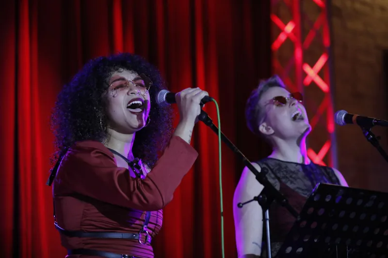 Two singers perform at microphones against a red velvet curtain backdrop, lit by purple and red stage lighting, with neon X…