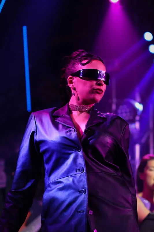 Performer on stage wearing mirrored sunglasses, sequined choker, and dark blazer, illuminated by purple and blue stage…